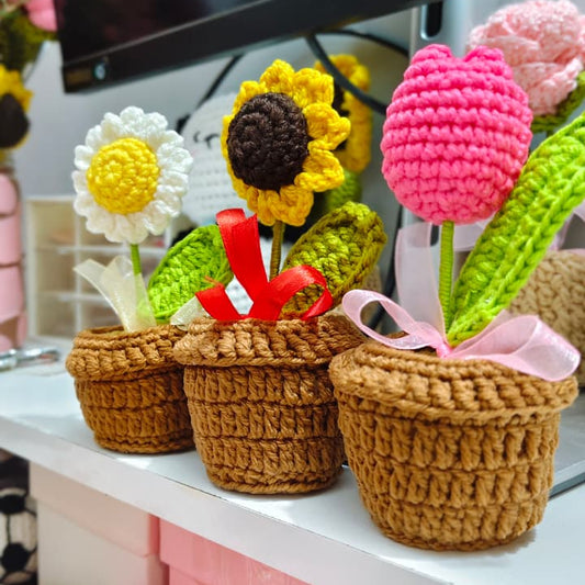 Three crocheted flower pots with colorful handmade flowers, including a sunflower, a daisy, and tulips, displayed on a shelf in front of a television.
