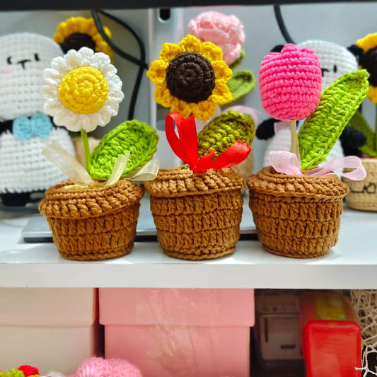Three crocheted potted sunflower and flower planties displayed on a white shelf in front of a black computer monitor, with more crocheted flowers visible in the foreground and background.