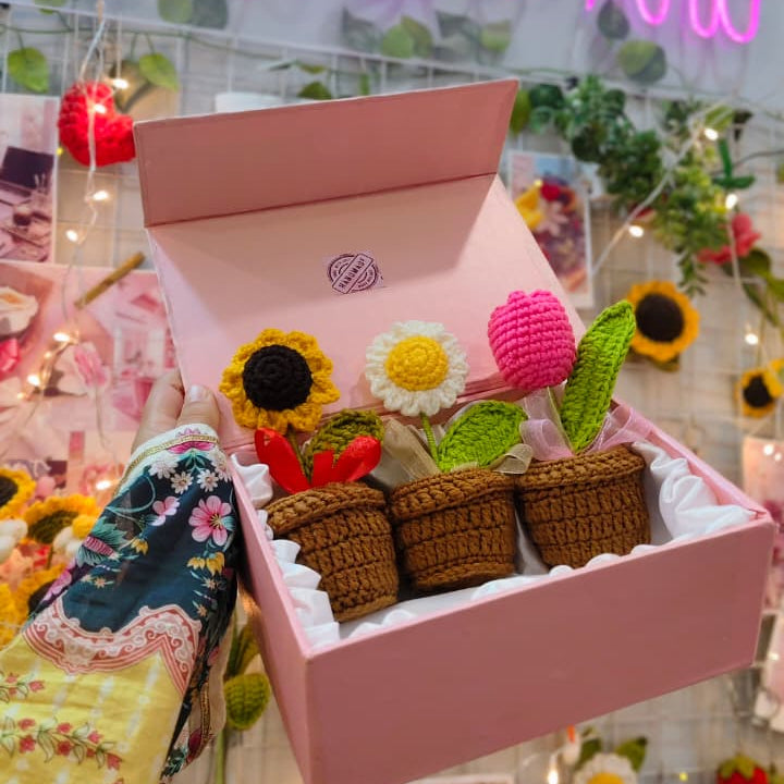 A pink gift box filled with crocheted flowers and small basket-like decorations, held by a hand wearing a floral-patterned sleeve. The background shows a shop interior with a pink neon sign reading 'Busy Bee Art' and decorative floral elements.