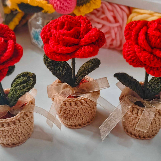 Three red crocheted roses in small brown pots on a white surface with colorful yarn in the background.