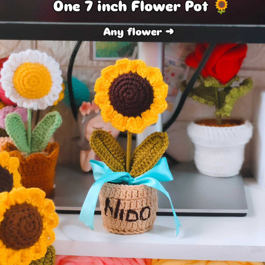 Crocheted sunflower in a pot with 'Nino' written on it, surrounded by other crocheted flowers.