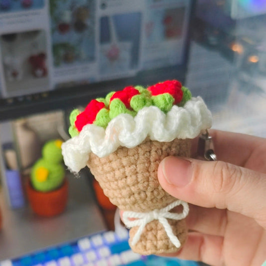 Hand holding a small crochet bouquet with red and green top against a blurred indoor background.