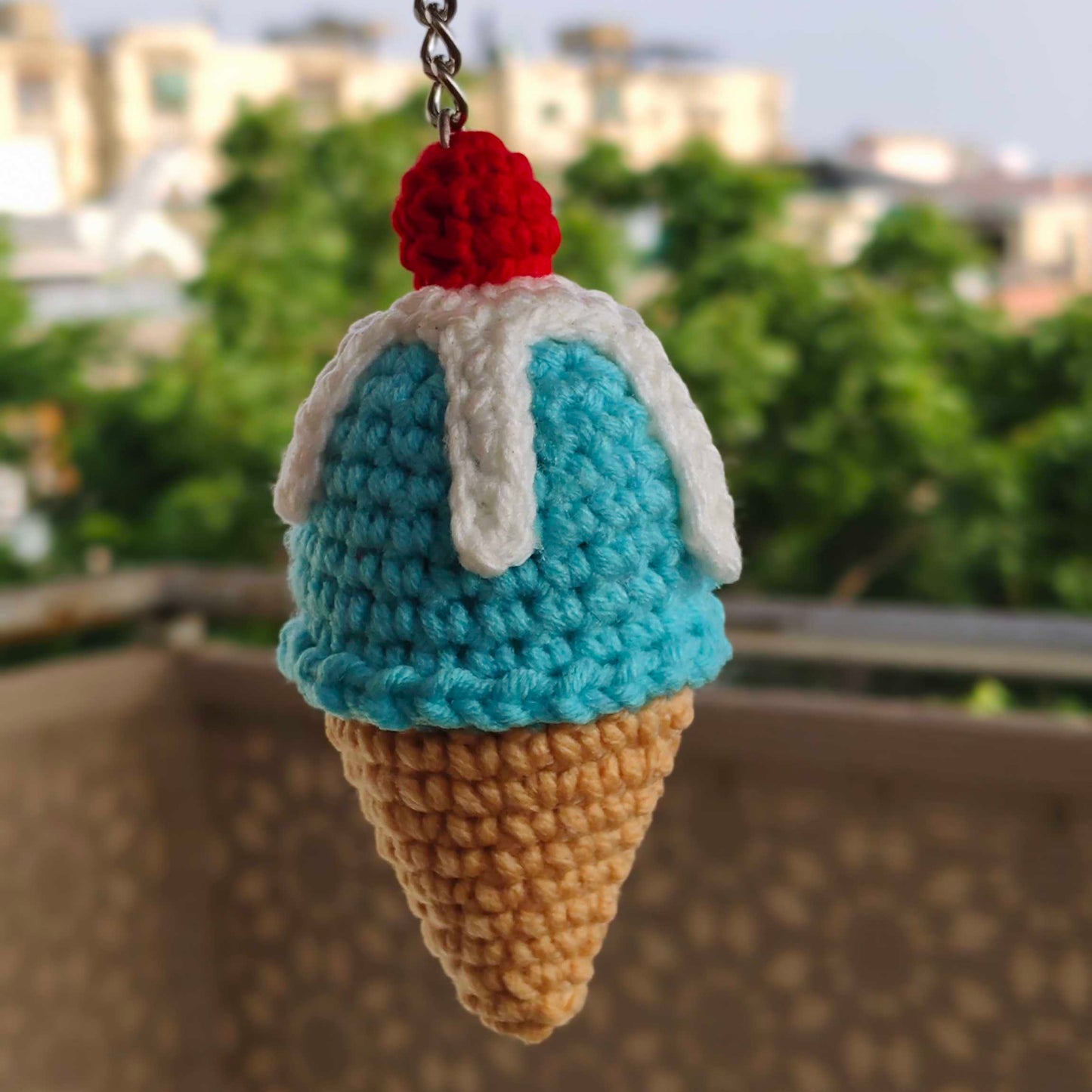 Crocheted ice cream cone keychain with a red cherry on top, held against a blurred outdoor background.