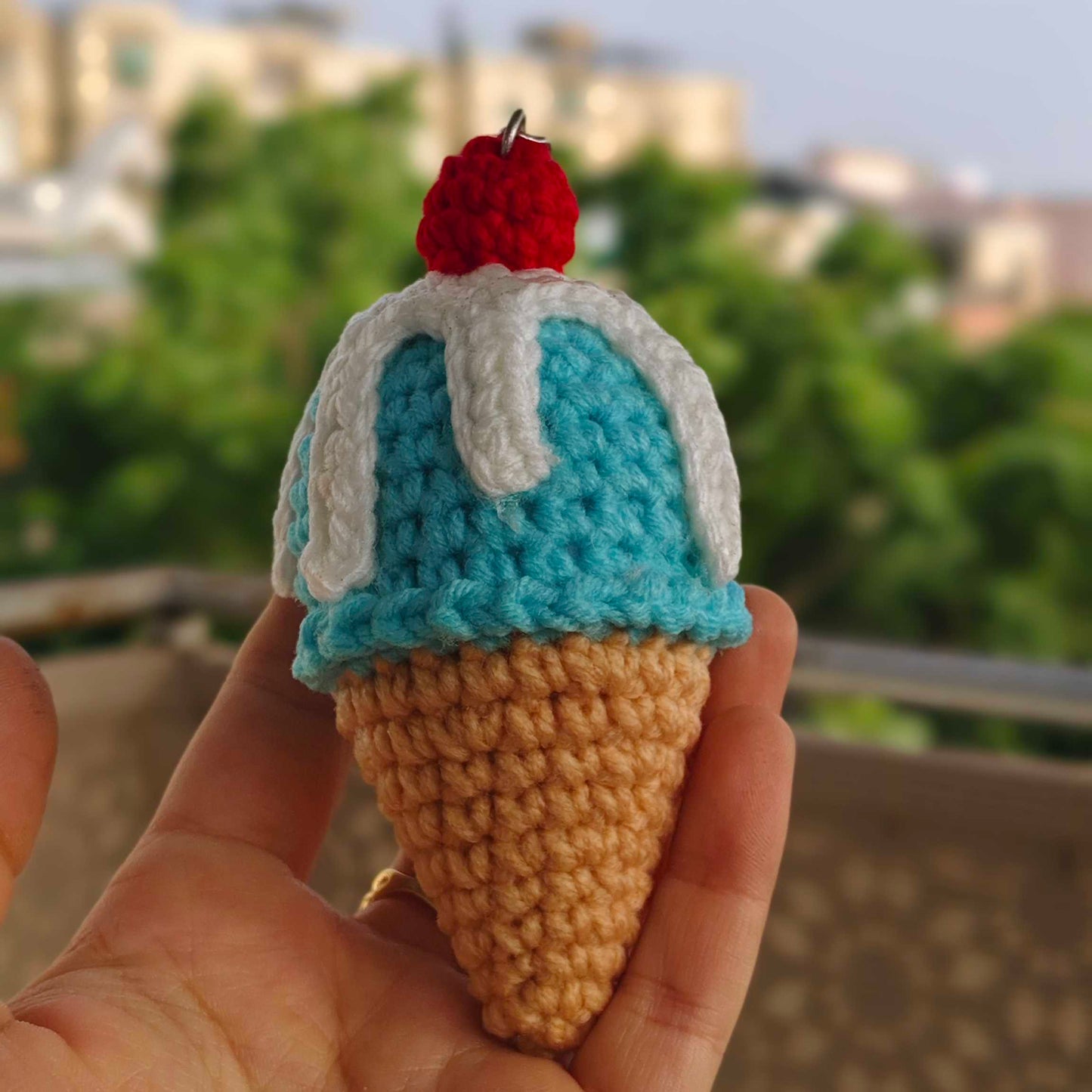 Hand holding a small crochet ice cream cone with a blue top and red cherry against a blurred outdoor background.