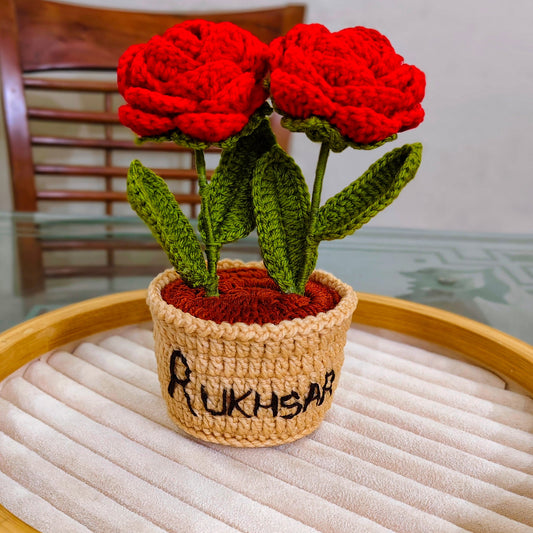 Crocheted red roses in a pot with 'Rukhsana' written on it, placed on a textured surface.