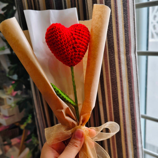 Red heart-shaped object on a green stem wrapped in brown paper with a ribbon, held by a hand against a striped curtain background.