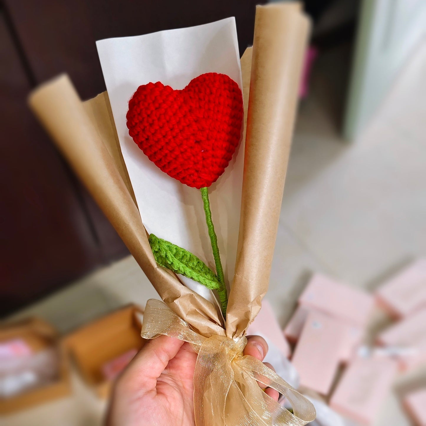 Bouquet of knitted red heart flowers wrapped in brown paper with a bow, held by a hand.