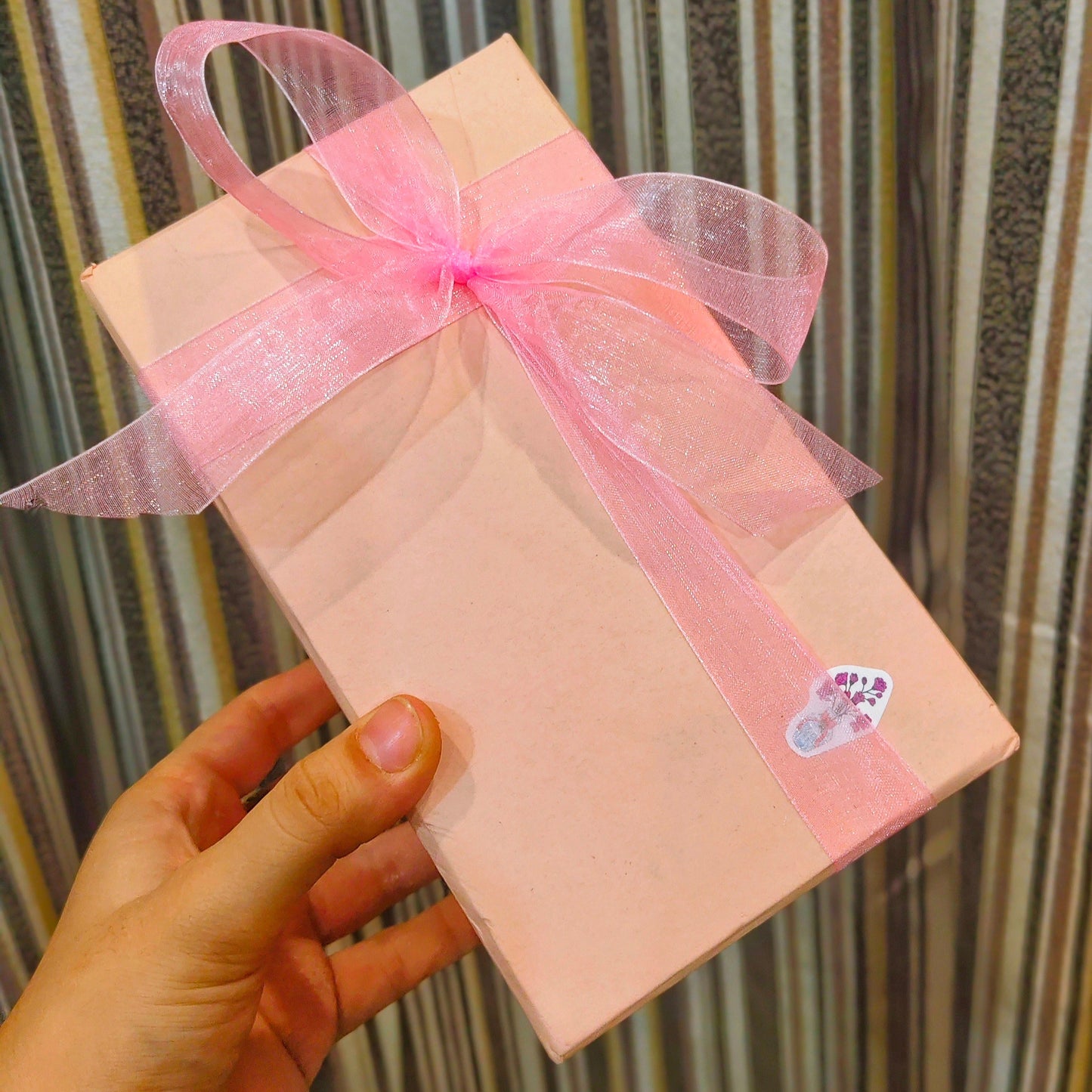 Pink gift box with a bow held by a hand against a striped curtain background