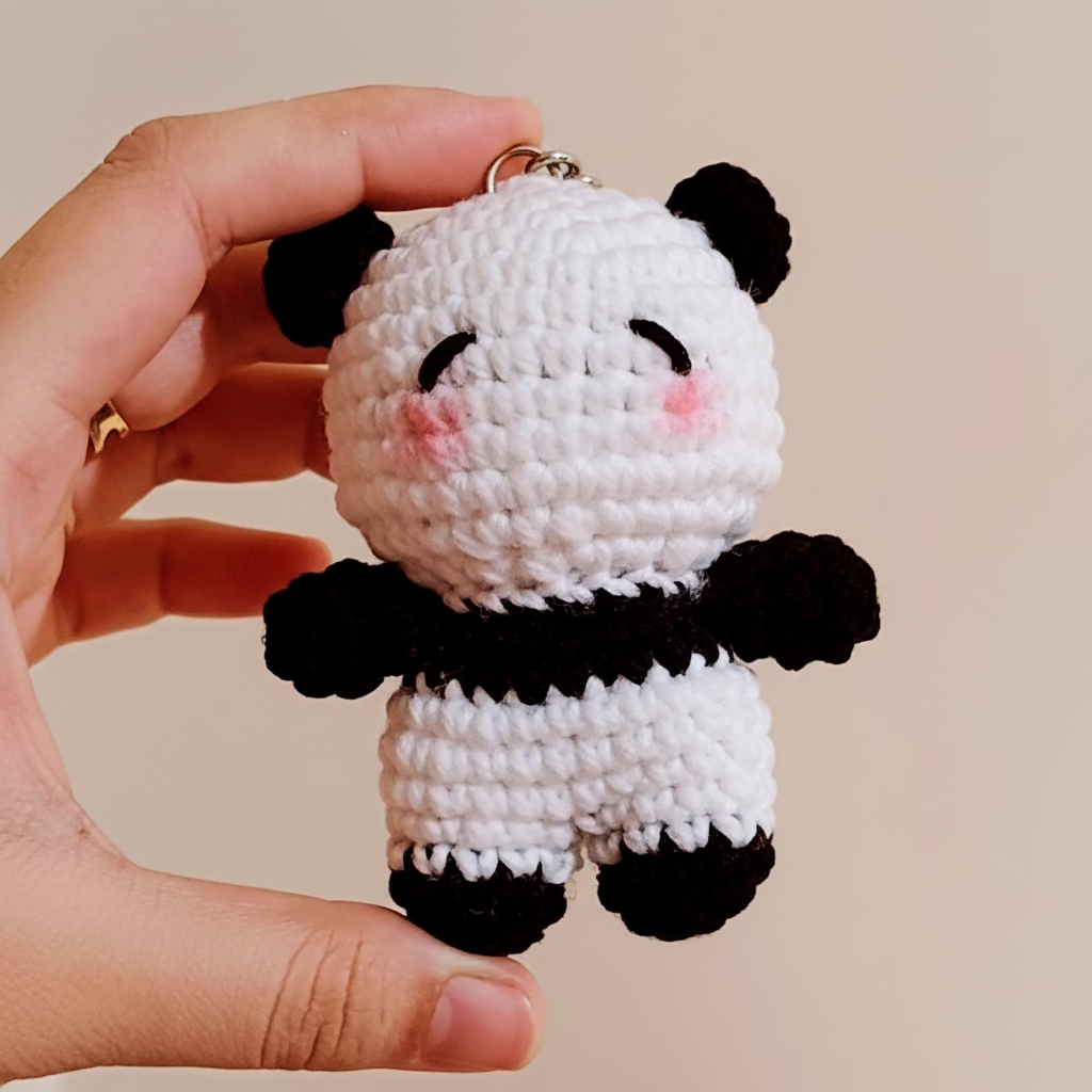 Hand holding a small crocheted panda keychain against a beige background