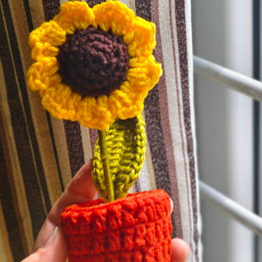 Handmade crochet sunflower in a pot held against a striped curtain.