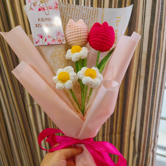 Bouquet of knitted flowers with a pink ribbon held by a person, against a striped curtain background.