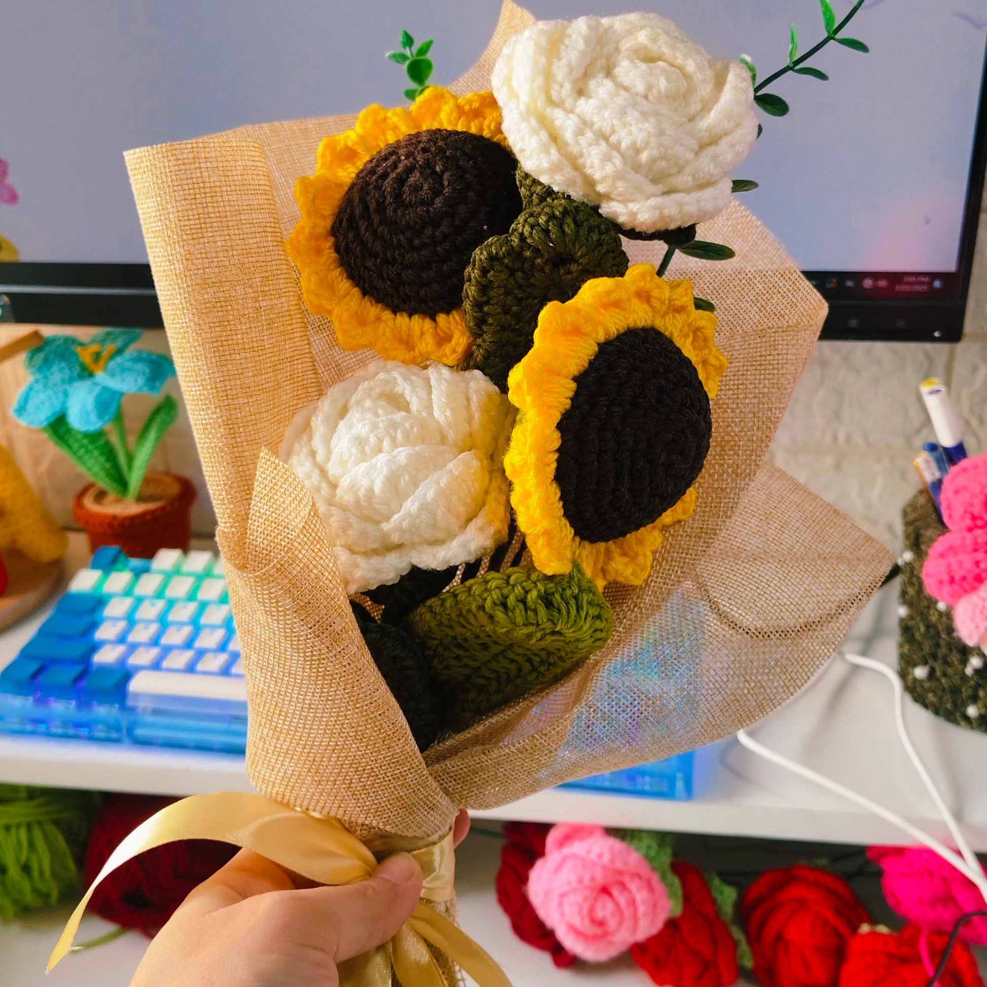 Hand holding a bouquet of knitted sunflowers and roses on a blurred indoor background