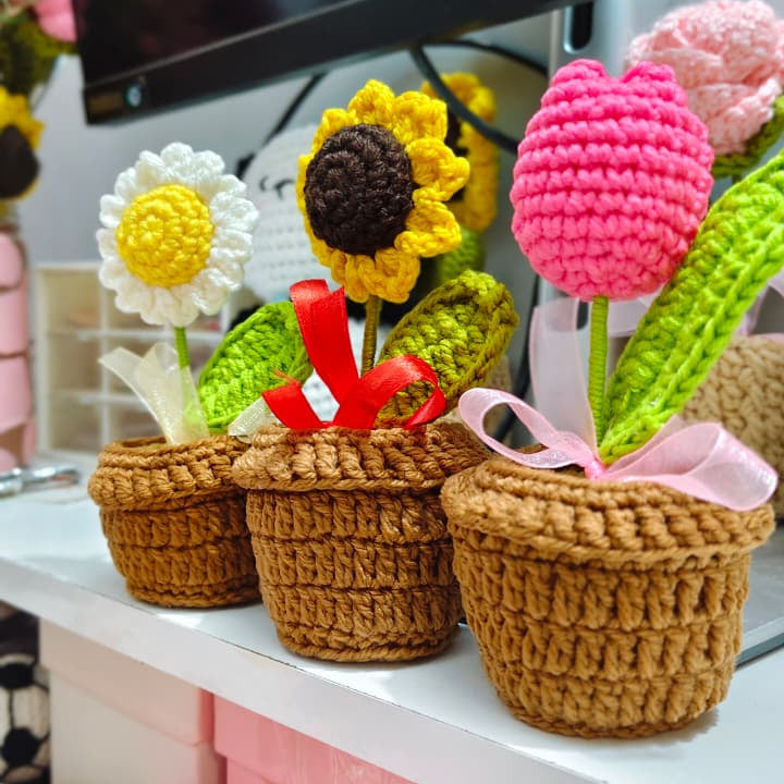 Three crocheted flower pots with colorful handmade flowers, including a sunflower, a daisy, and tulips, displayed on a shelf in front of a television.