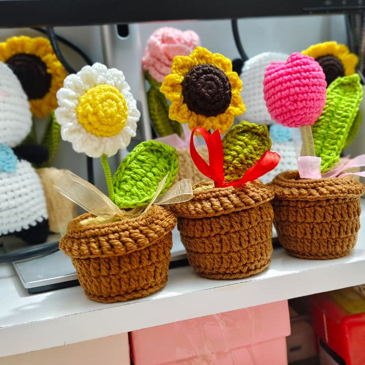A collection of handmade crocheted sunflower and flower pot ornaments displayed on a white shelf in front of a black computer monitor.