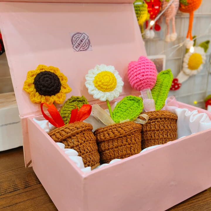 A pink crocheted flower gift box filled with handmade crochet flowers and small brown pots, displayed on a wooden surface with other colorful crochet items in the background.