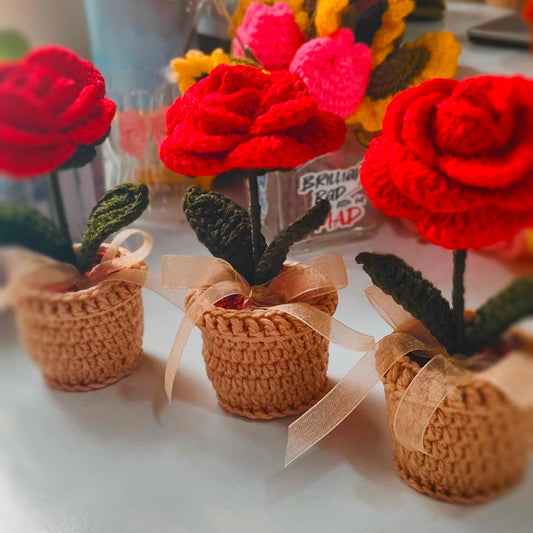 Three crocheted red roses in small brown pots on a white surface.
