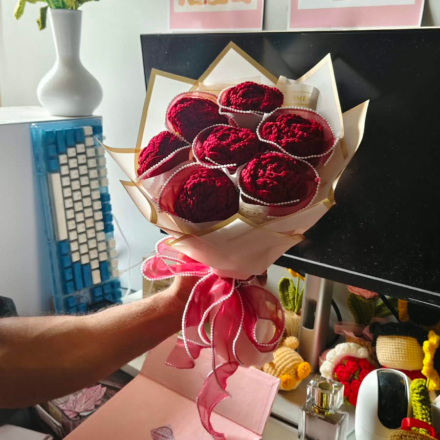 A hand holds a bouquet of red roses wrapped in white paper with a pink ribbon, in front of a desk with a monitor, keyboard, and framed art.