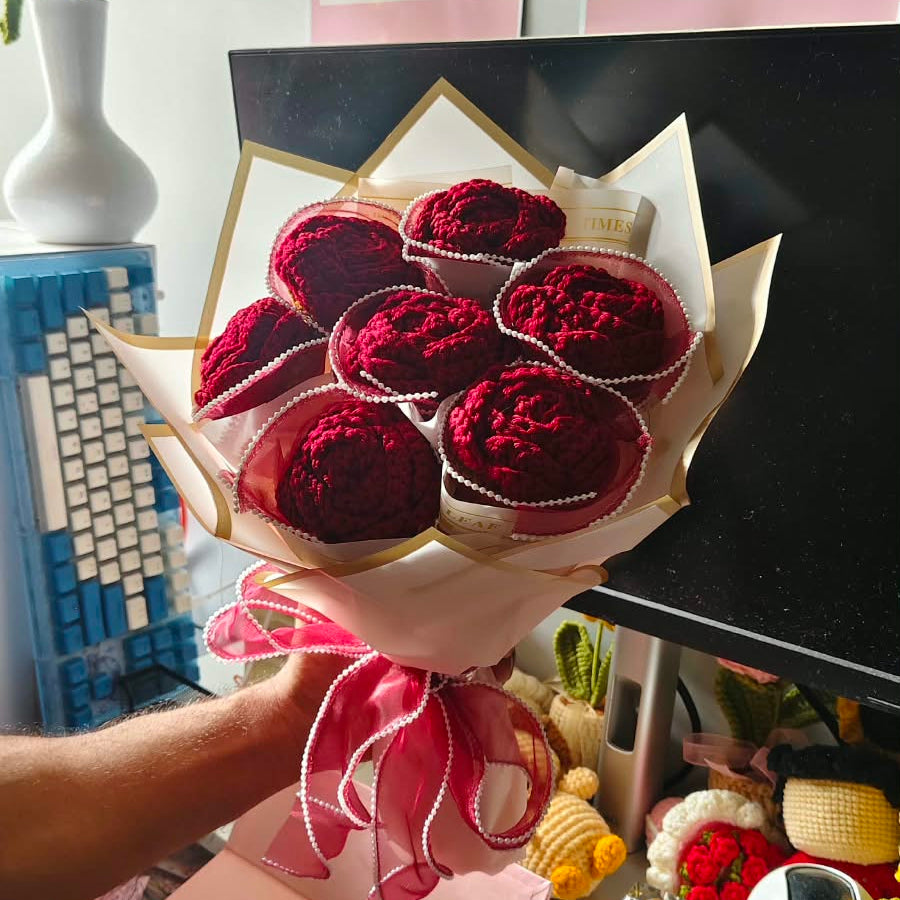 A hand holding a bouquet made of red velvet-covered heart-shaped chocolates wrapped in pink paper with a pink ribbon, set against a busy desk background with a computer monitor and framed posters.