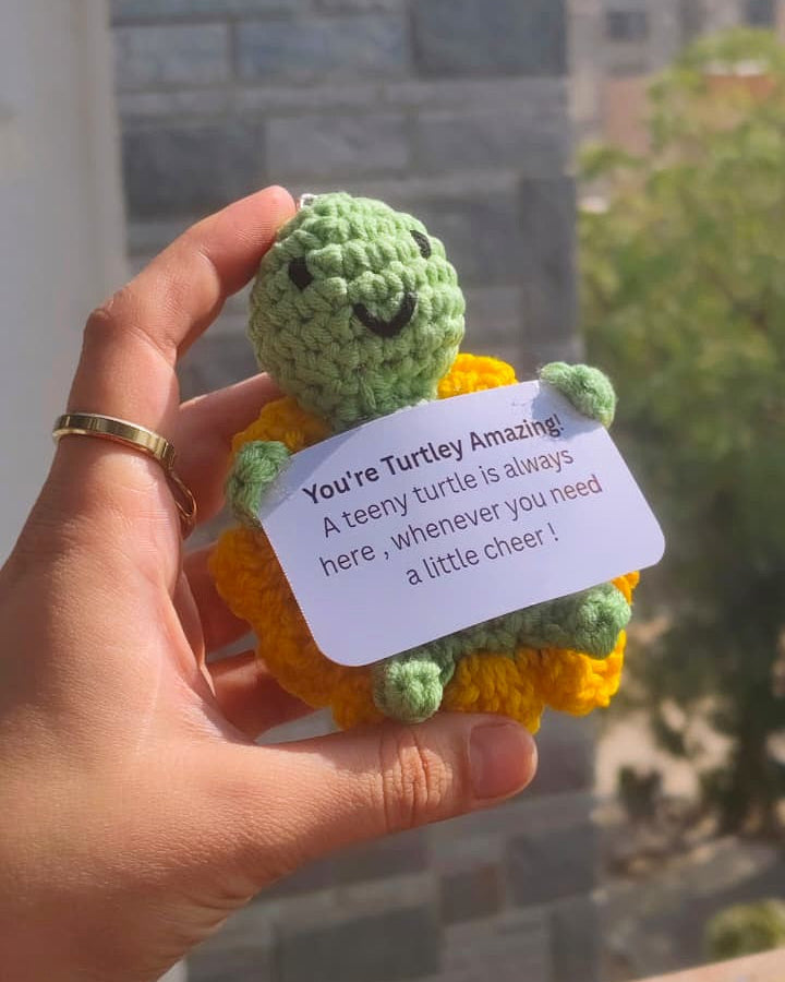 Hand holding a small green and orange crochet turtle with a message card, outdoors.