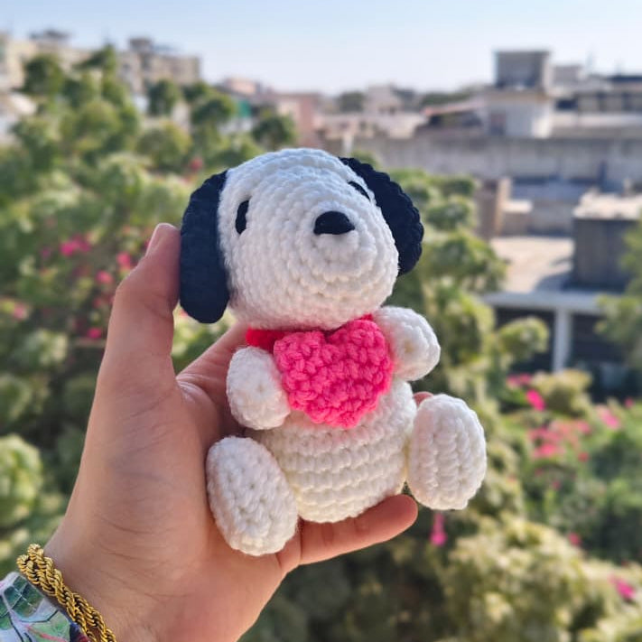 Hand holding a small crocheted dog toy with a pink heart against a blurred outdoor background.