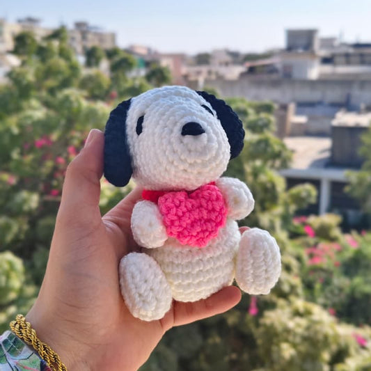 Hand holding a small crocheted dog toy with a pink heart against a blurred outdoor background.