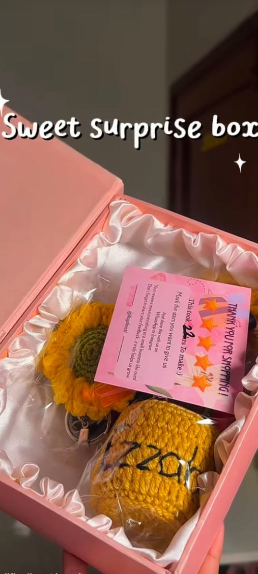 A pink gift box with a white satin lining, containing a yellow sunflower, a pink handwritten note, and a yellow knitted item. The photo is taken in a real indoor setting and includes social media interface overlays.