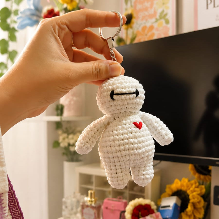 A hand holds a small crocheted white character keychain with a red heart on its chest, set against a cozy, decorated room background with a TV, flowers, and shelves.
