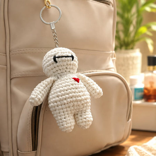 A cream crocheted plush keychain character with a small red heart and black stitched eyes hangs from a silver key ring on a beige backpack, set on a wooden table with a blurred background.
