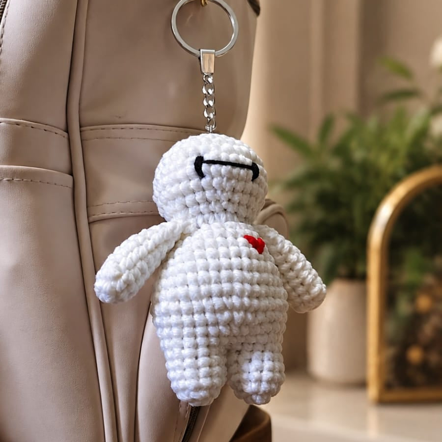 A white crocheted character keychain with a small red heart and black stitched eyes, hanging from a beige bag in an indoor setting.
