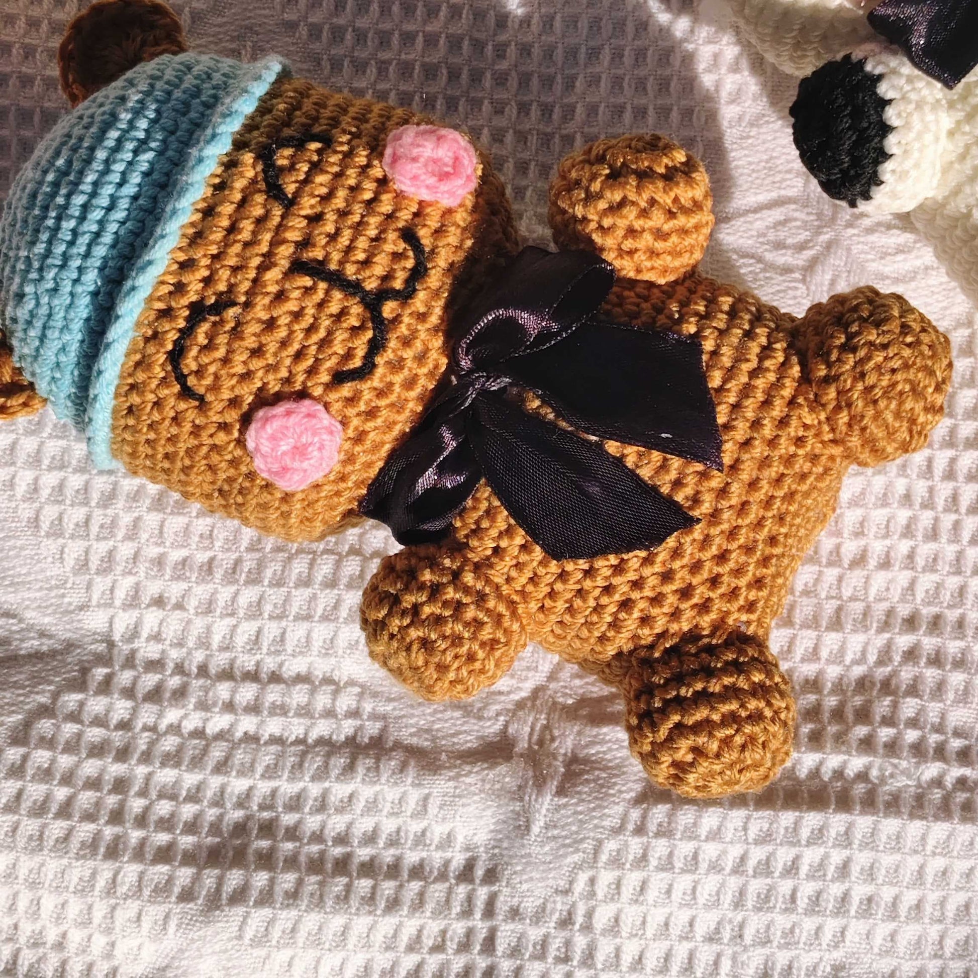 Crocheted bear toy with a blue beanie and black bow tie on a textured white surface.