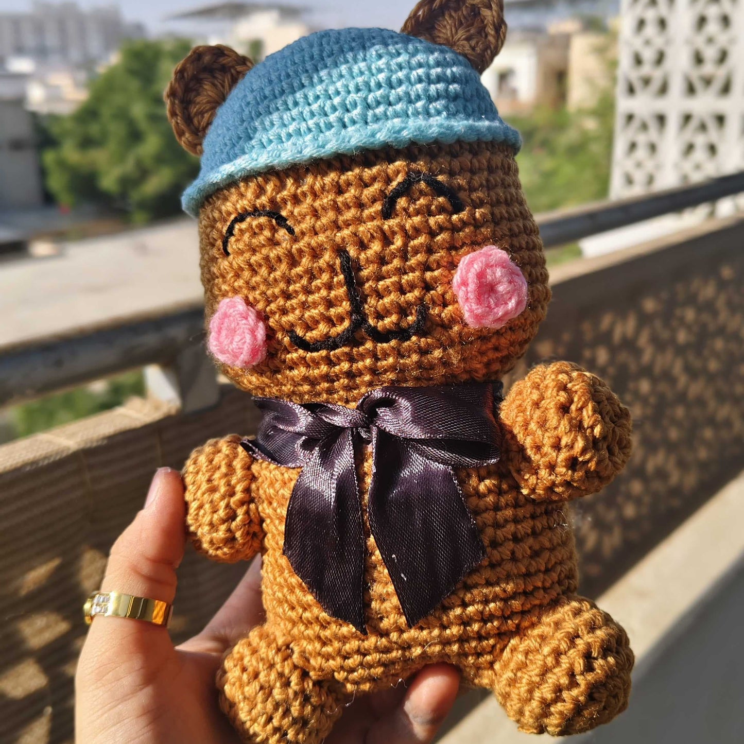 Hand holding a small brown teddy bear with a blue hat and pink cheeks, outdoors.