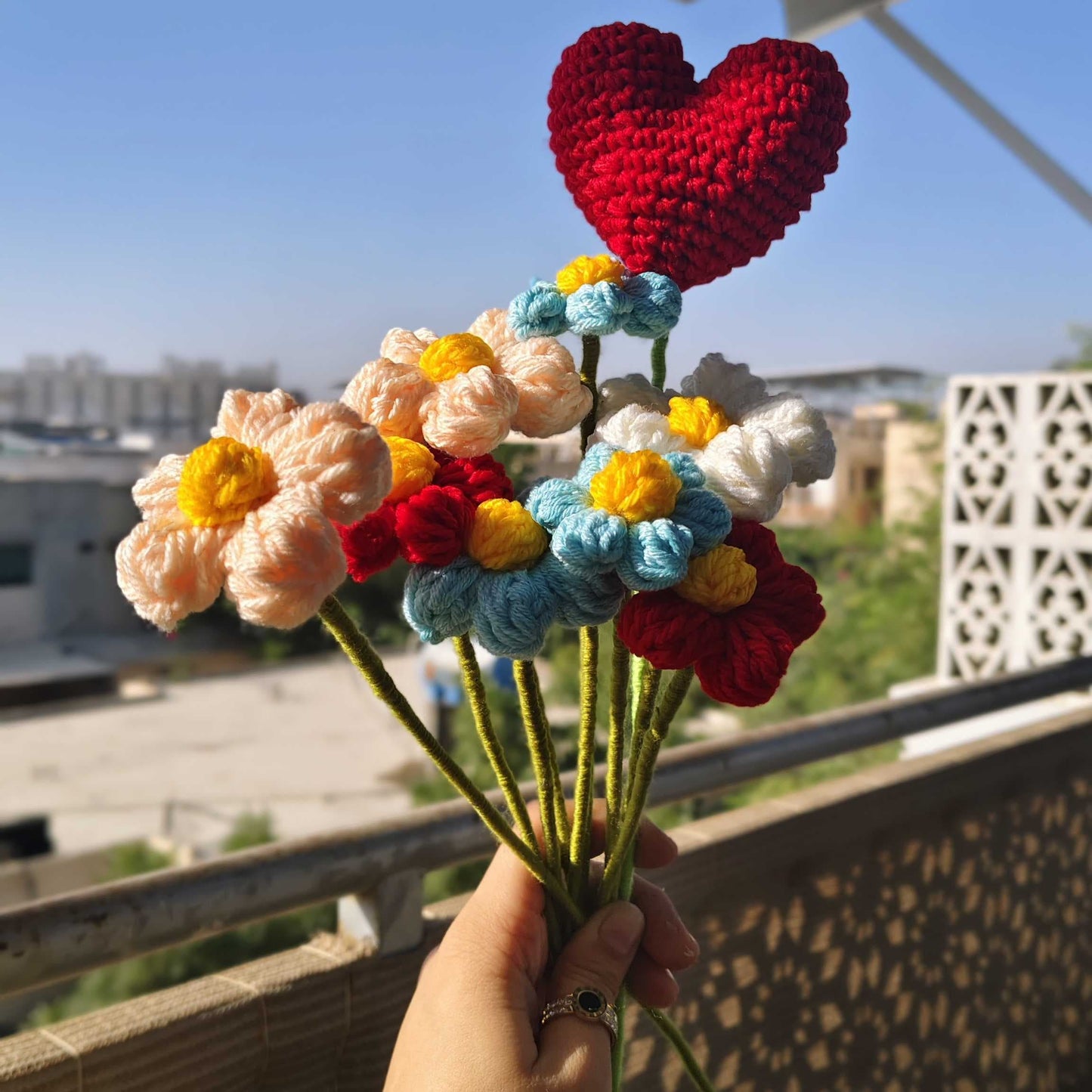 Hand holding a bouquet of crocheted flowers with a red heart on top, under a solar panel.