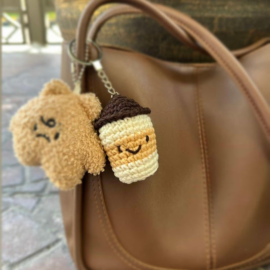 Two crochet keychains, one resembling a bear and the other a coffee cup, attached to a brown leather bag.