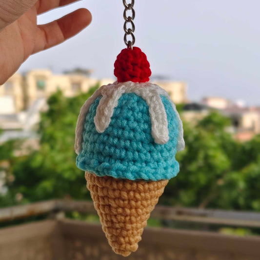 Crocheted ice cream cone keychain with a red cherry on top held by a hand against a blurred outdoor background.