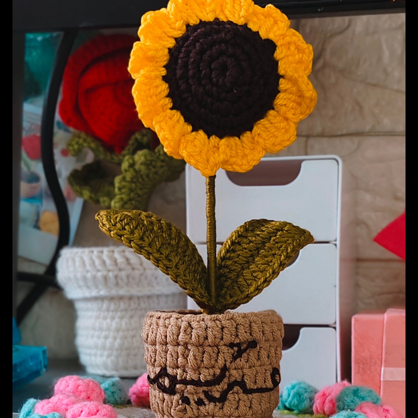 Crocheted sunflower in a pot with colorful yarn balls on a table.
