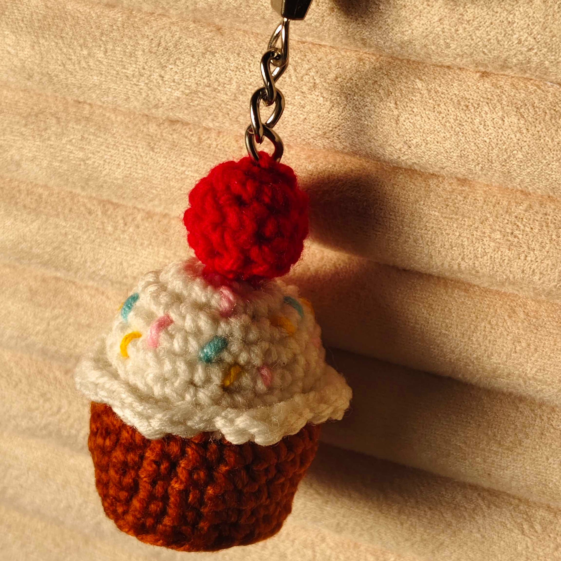 Crocheted cupcake keychain with a red cherry on top against a beige background