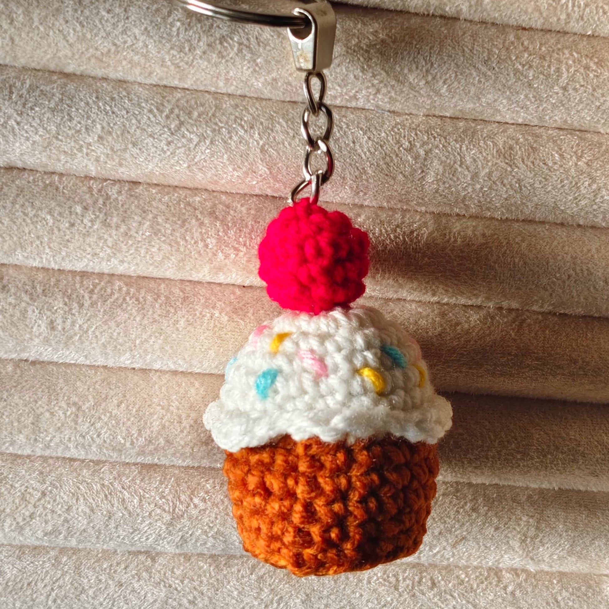 Crocheted cupcake keychain with a red cherry on top against a textured beige background