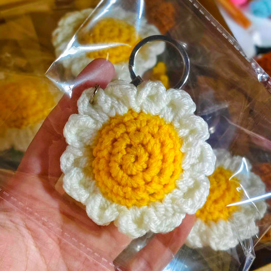 Hand holding a crocheted keychain with a yellow center and white petals, with packaging in the background.