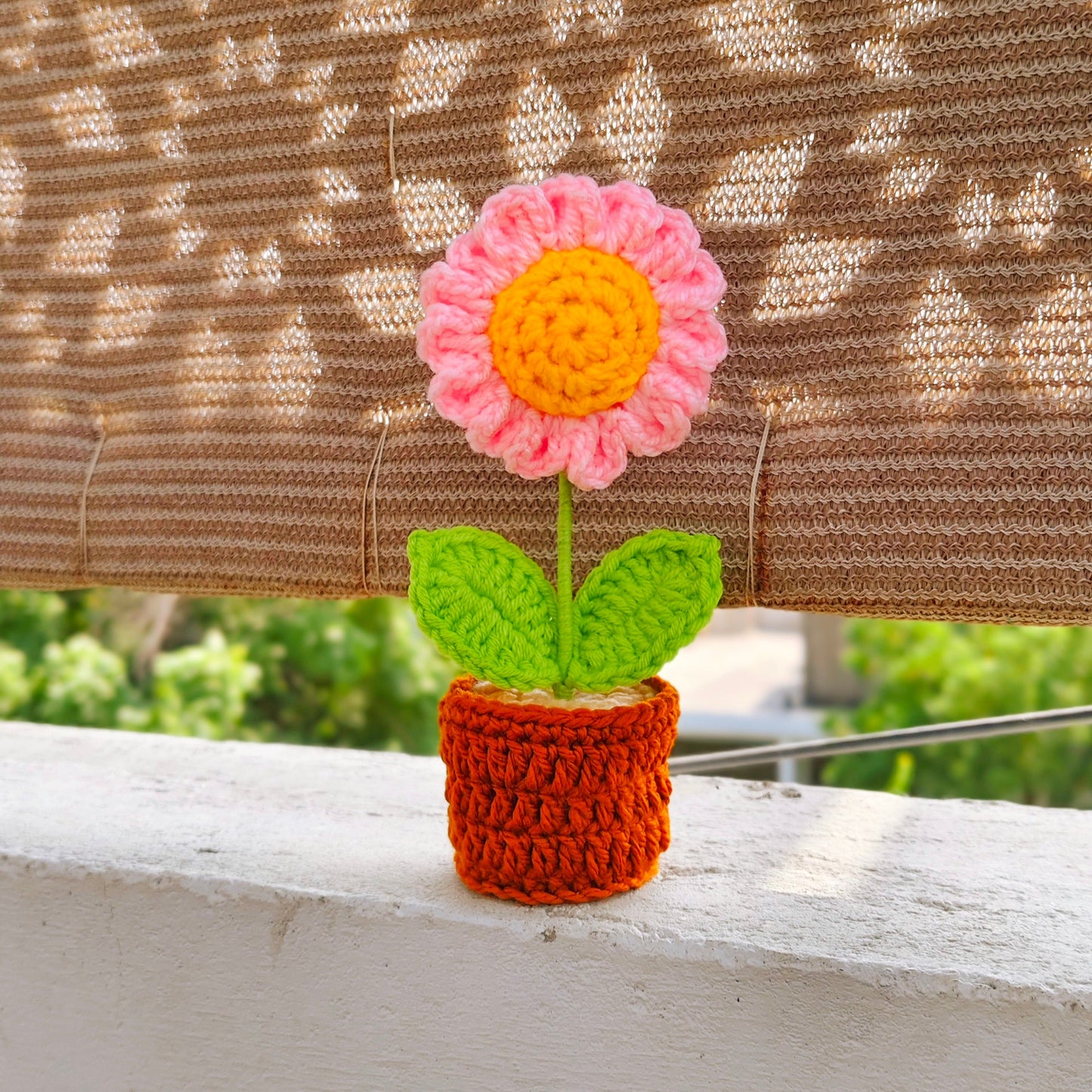 Crocheted flower in a pot on a wooden surface with a patterned curtain in the background.