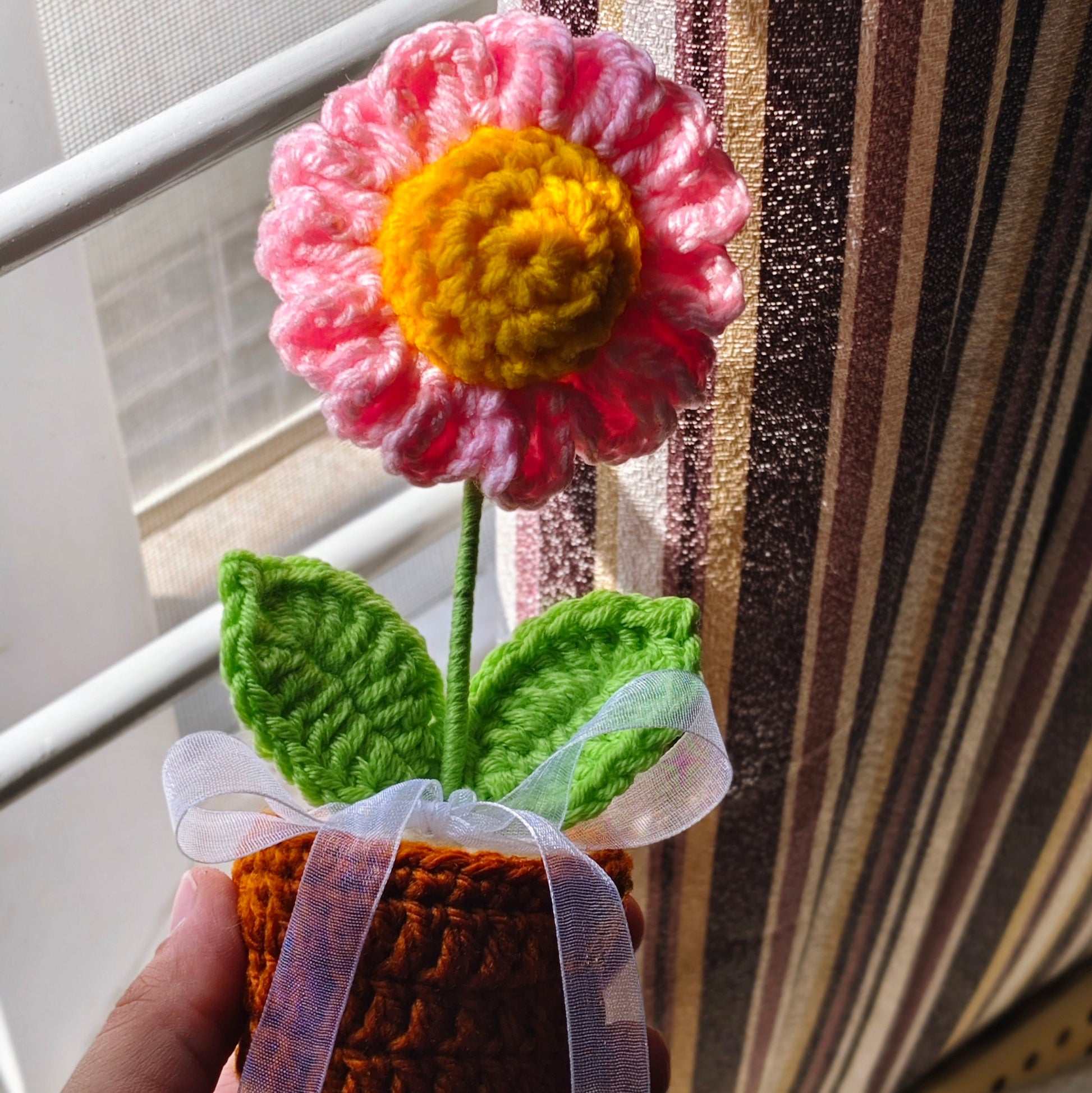 Handmade crochet daisy  flower with pink petals and yellow center held against a striped background.