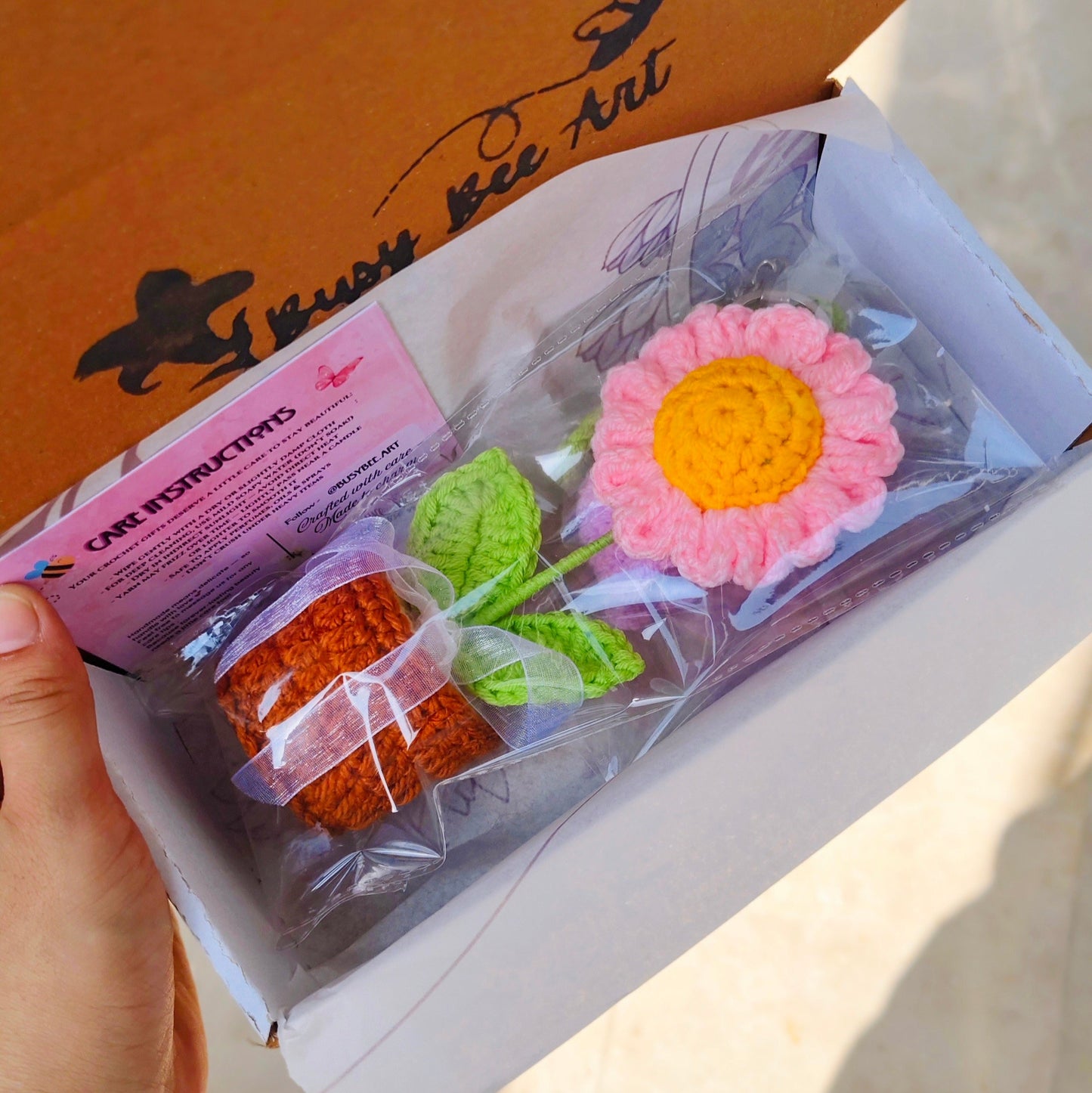 Hand holding a box with crochet daisy flowers and leaves inside, against a blurred background.