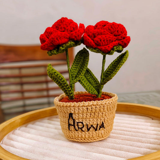 Crocheted red flowers in a brown pot with 'Arwa' written on it, placed on a wooden surface.