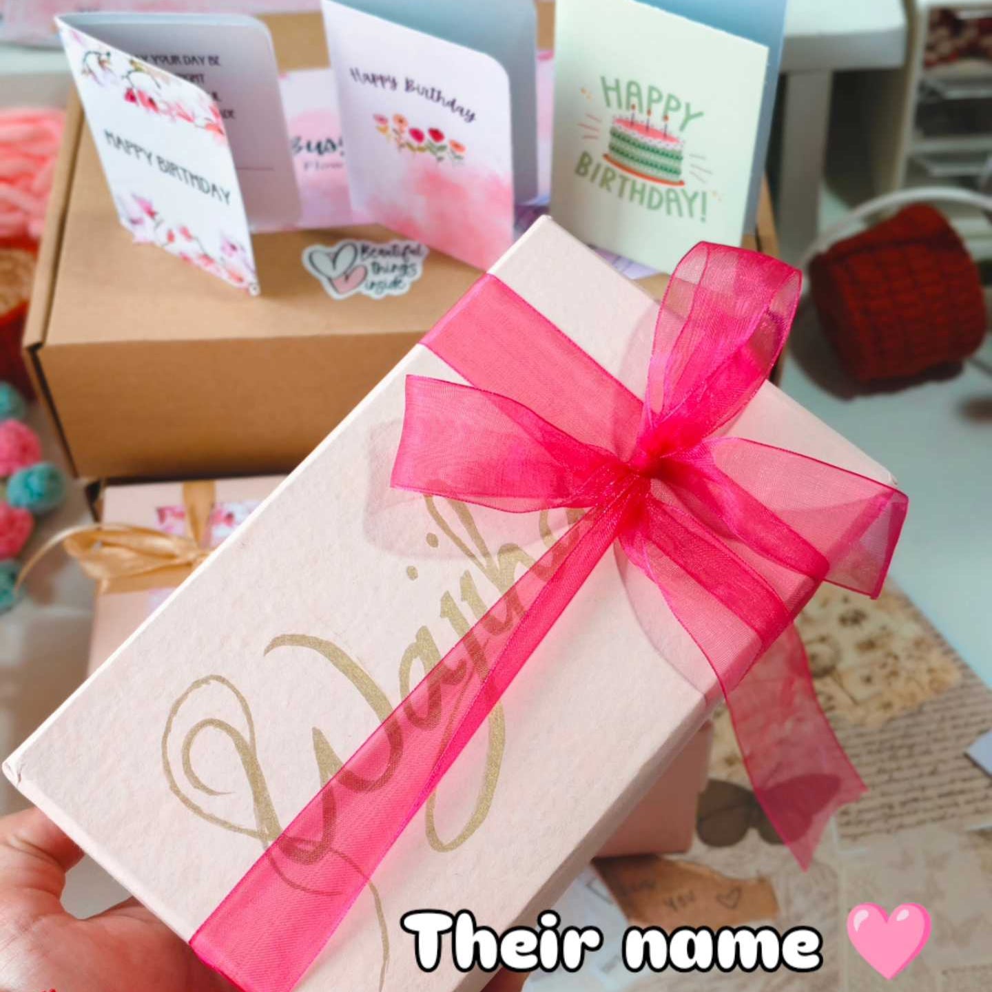 Gift box with a pink ribbon and personalized name on a textured surface with greeting cards in the background.