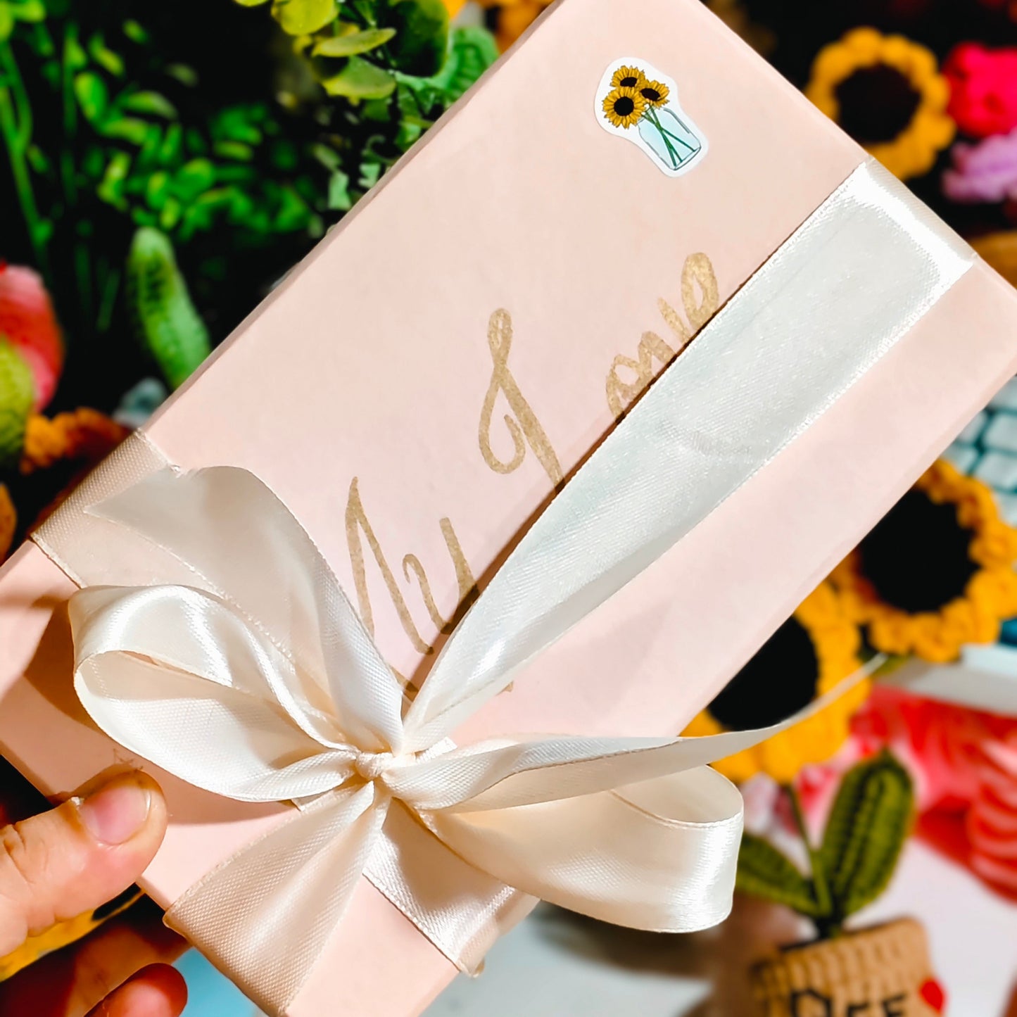 Gift box with a white ribbon held by a hand, surrounded by sunflowers and decorative items.