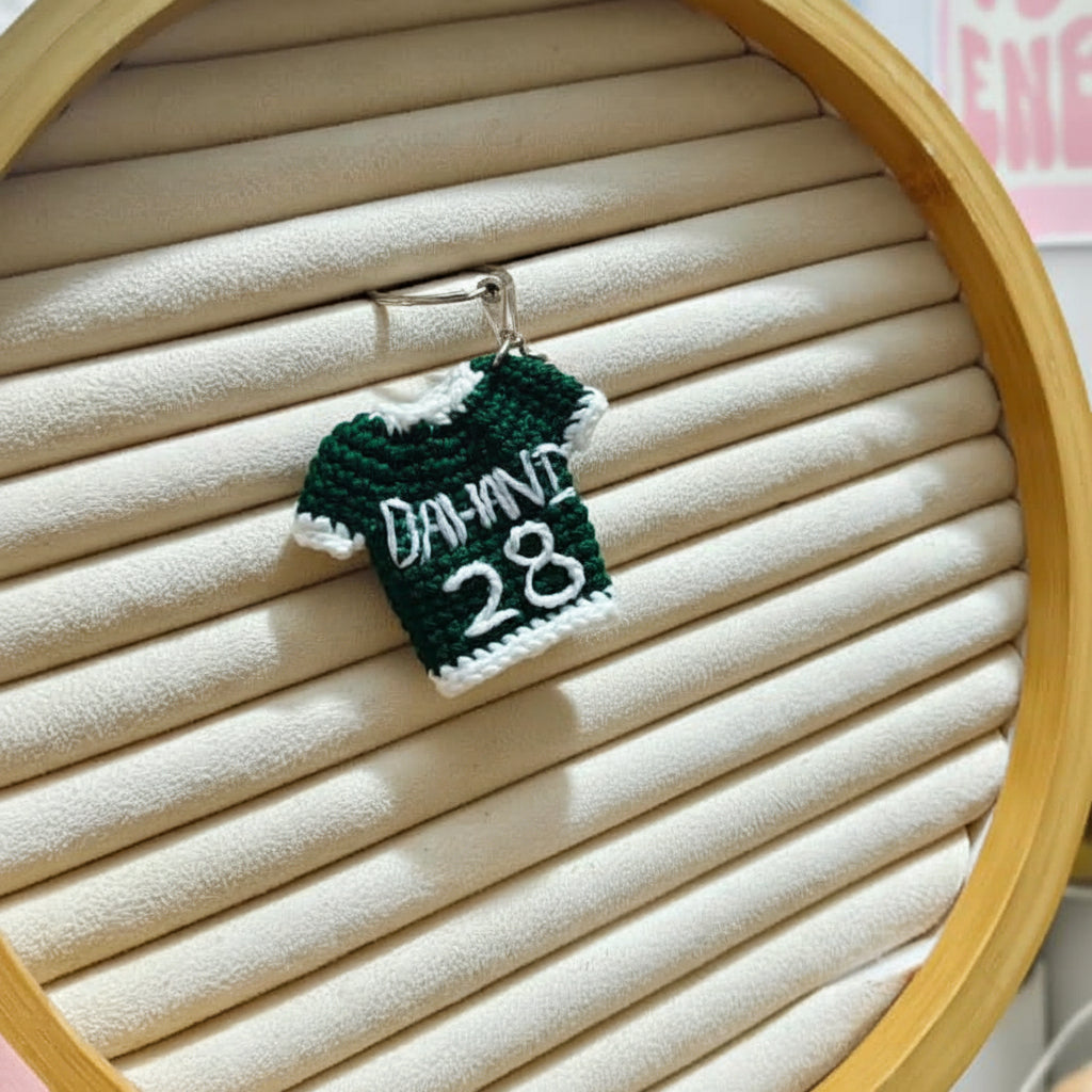 A miniature crochet keychain featuring a green and white sports jersey with the number 28 and a red-and-beige baseball, displayed inside a wooden hoop on a beige slatted backing.