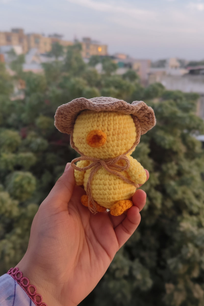 Hand holding a small crochet duck with a hat against a blurred outdoor background