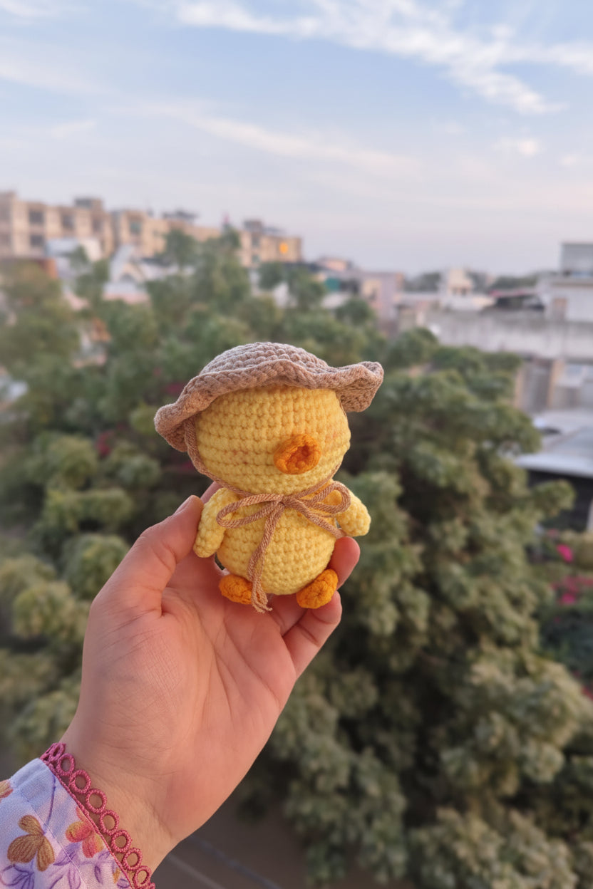 Hand holding a small yellow crocheted duck toy with a hat against a cityscape background.