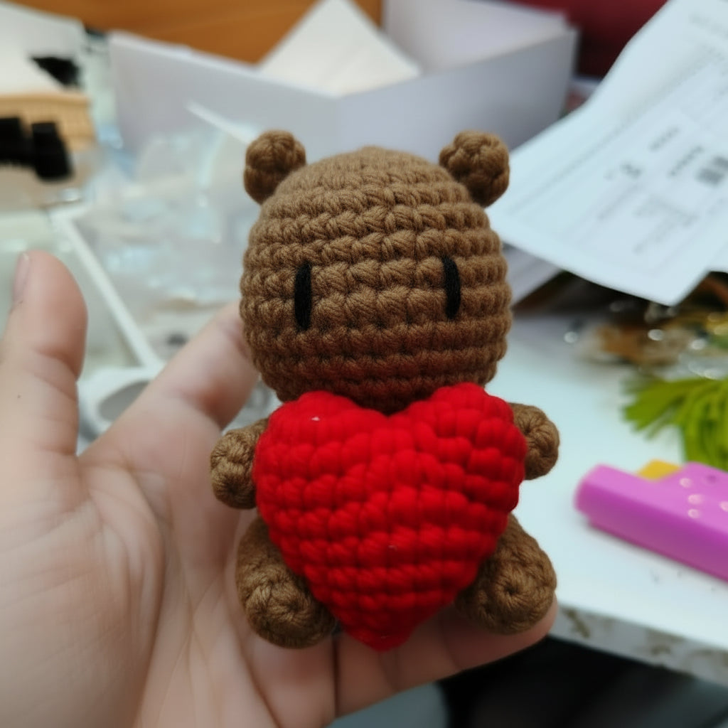 A hand holds a small crocheted white bear toy holding a bright red heart-shaped pillow, with a cluttered desk background in the blurred distance.