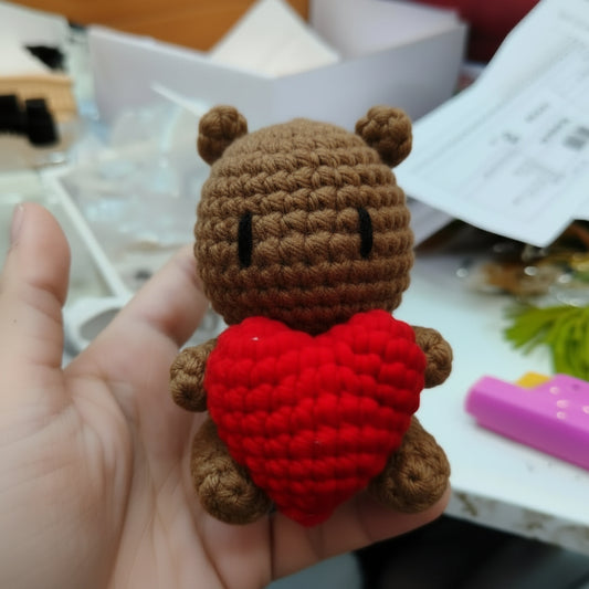 A hand holds a small crocheted white bear toy holding a bright red heart-shaped pillow, with a cluttered desk background in the blurred distance.
