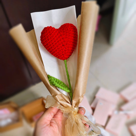 Bouquet of knitted red heart flowers wrapped in brown paper with a bow, held by a hand.